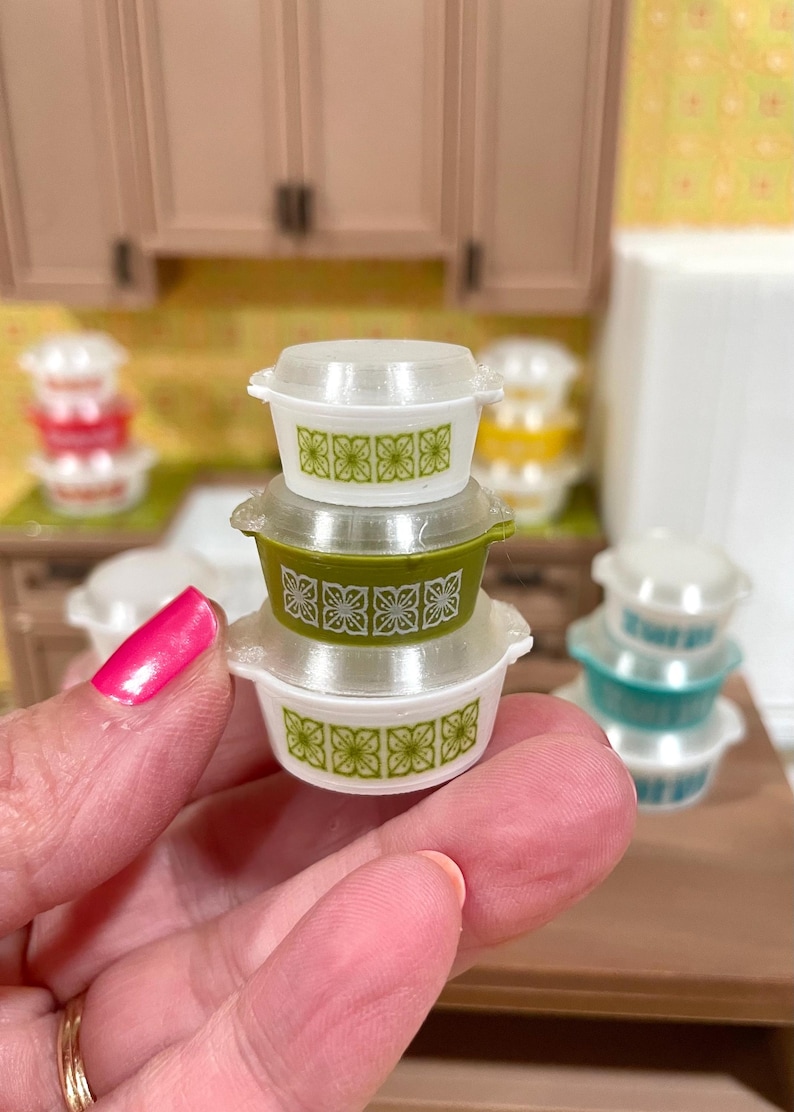 May include: A stack of three miniature white and green casserole dishes with clear lids. Each dish features a repeating green floral pattern. The dishes are held in a person's hand, with other similar dishes in the background.