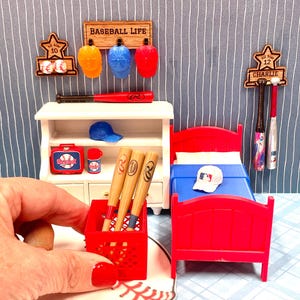May include: A miniature baseball-themed bedroom set featuring a white hutch, a red bed with blue bedding, and a red crate holding three wooden bats. The display includes wall-mounted wooden racks labeled BASEBALL LIFE, ALL STAR 10, and CHARLIE.