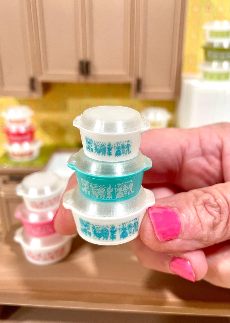 May include: A stack of three miniature Pyrex-style bowls with lids. The bowls are white and teal, featuring a teal rooster and floral design. The bowls are held in a hand.