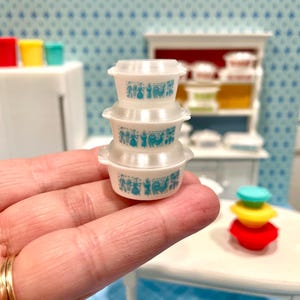 May include: Miniature white Pyrex-style bowls with lids, stacked in a set of three. Each bowl features a blue floral and animal design. The bowls are part of a dollhouse kitchen scene, with other colorful miniature kitchenware in the background.