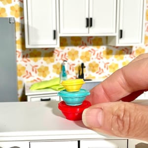 May include: A set of three nesting bowls in red, blue, and yellow. The bowls are stacked on top of each other on a white countertop.