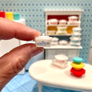 May include: A miniature white casserole dish with a lid, adorned with a blue floral pattern. The tiny dish is held in a hand, showcasing its small size. Other miniature kitchen items are in the background.
