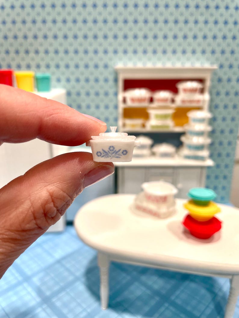 May include: A miniature white casserole dish with a lid, adorned with a blue floral pattern. The tiny dish is held in a hand, showcasing its small size. Other miniature kitchen items are in the background.