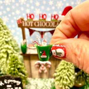 May include: A miniature green mug with a Santa hat design, filled with a miniature snowman, candy cane, and a silver stirrer. The mug is being held by a hand with a snowman nail design. The background features a miniature Christmas village with a sign that reads "Ho Ho Ho Hot Chocolate".