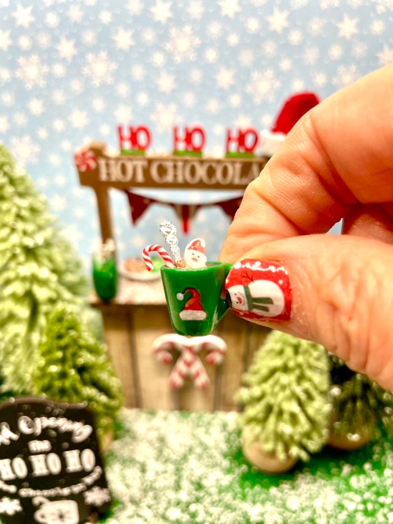 May include: A miniature green mug with a Santa hat design, filled with a miniature snowman, candy cane, and a silver stirrer. The mug is being held by a hand with a snowman nail design. The background features a miniature Christmas village with a sign that reads "Ho Ho Ho Hot Chocolate".