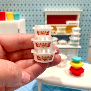 May include: A stack of three miniature white plastic bowls with red and yellow floral designs. The bowls are held in a hand, with a miniature kitchen scene in the background. Other colorful miniature kitchen items are visible.