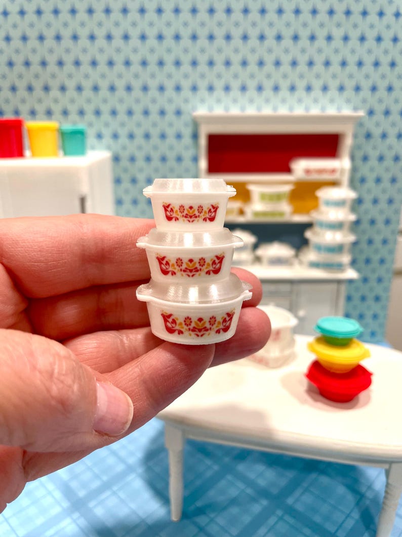 May include: A stack of three miniature white plastic bowls with red and yellow floral designs. The bowls are held in a hand, with a miniature kitchen scene in the background. Other colorful miniature kitchen items are visible.