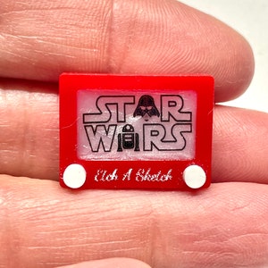 May include: Red plastic toy resembling a classic Etch A Sketch with the words "STAR WARS" and a Darth Vader silhouette on the screen. The words "etch a sketch" are printed below the screen.