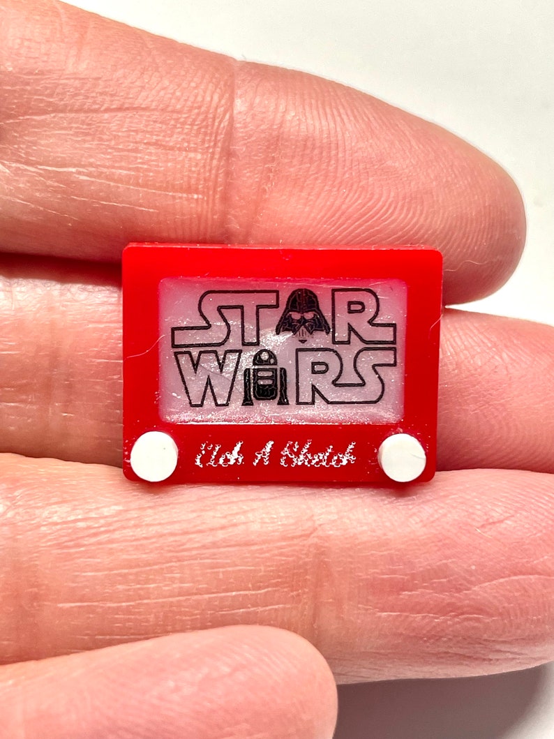 May include: Red plastic toy resembling a classic Etch A Sketch with the words "STAR WARS" and a Darth Vader silhouette on the screen. The words "etch a sketch" are printed below the screen.