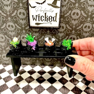 Puede incluir: Cuatro calderos negros en miniatura con purpurina de diferentes colores y detalles de temática de Halloween en un estante negro. El estante está sobre un suelo de cuadros negros y blancos. Un cartel enmarcado sobre el estante dice "Perfectly Wicked".