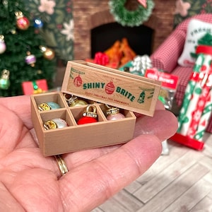 May include: Miniature wooden box of Shiny Brite glass Christmas tree ornaments. The box is open, revealing six colorful ornaments. The box has a vintage design with the words "Shiny Brite" printed on the front. The ornaments are in various colors, including red, gold, and pink.