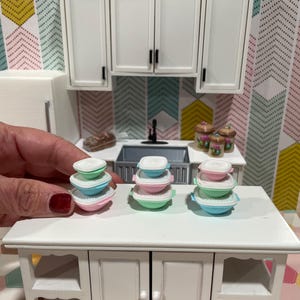 May include: Miniature kitchen scene featuring a white cabinet with stacked, colorful food containers. The containers are pastel pink, blue, and green with white lids. A white cabinet and a patterned wallpaper are in the background.