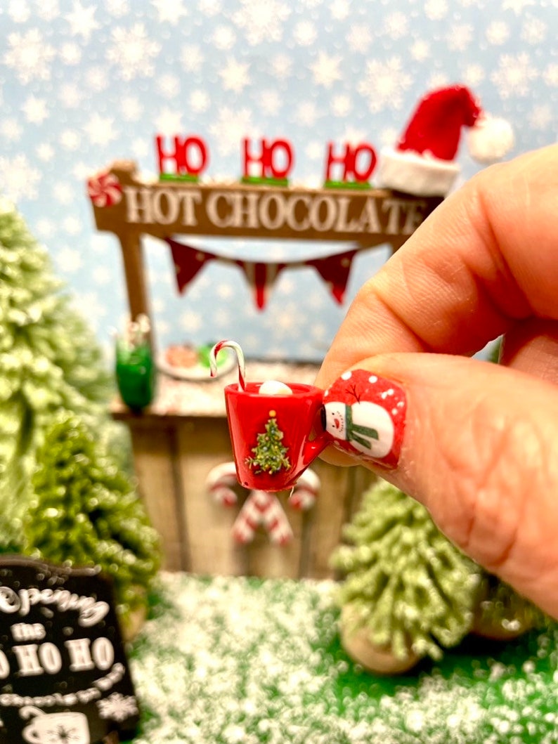 May include: A miniature red mug with a green Christmas tree design and a candy cane stirrer. The mug is filled with white marshmallows and is being held by a hand with red and white snowman nail art.