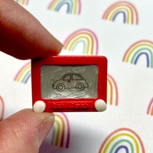 May include: A miniature red Etch A Sketch toy with a white car drawing on the screen. The toy has white knobs on the sides and the text "Etch A Sketch" is visible on the bottom.
