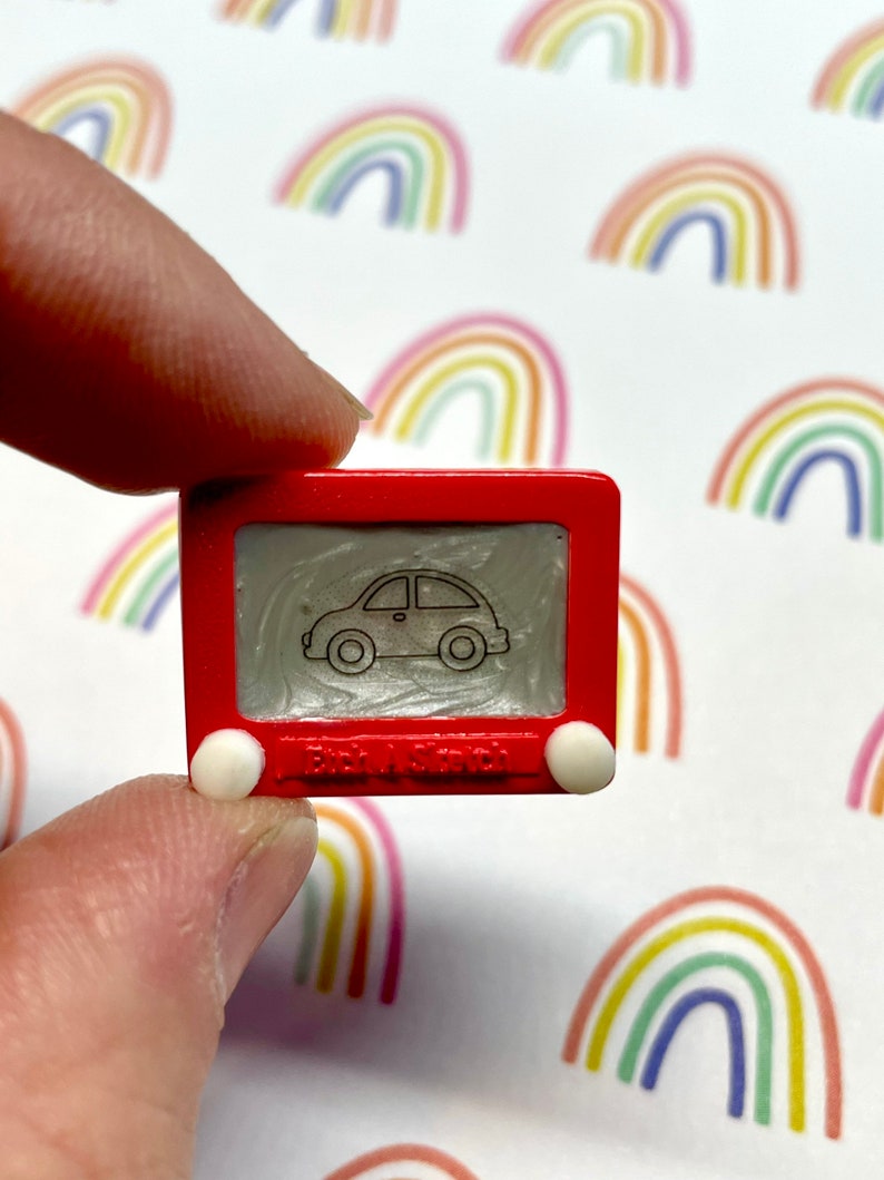 May include: A miniature red Etch A Sketch toy with a white car drawing on the screen. The toy has white knobs on the sides and the text "Etch A Sketch" is visible on the bottom.
