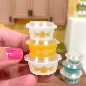 May include: A stack of miniature Pyrex-style bowls. The stack includes a yellow bowl, two white bowls, and clear lids. Each bowl features a floral design in yellow. Other bowls are visible in the background.