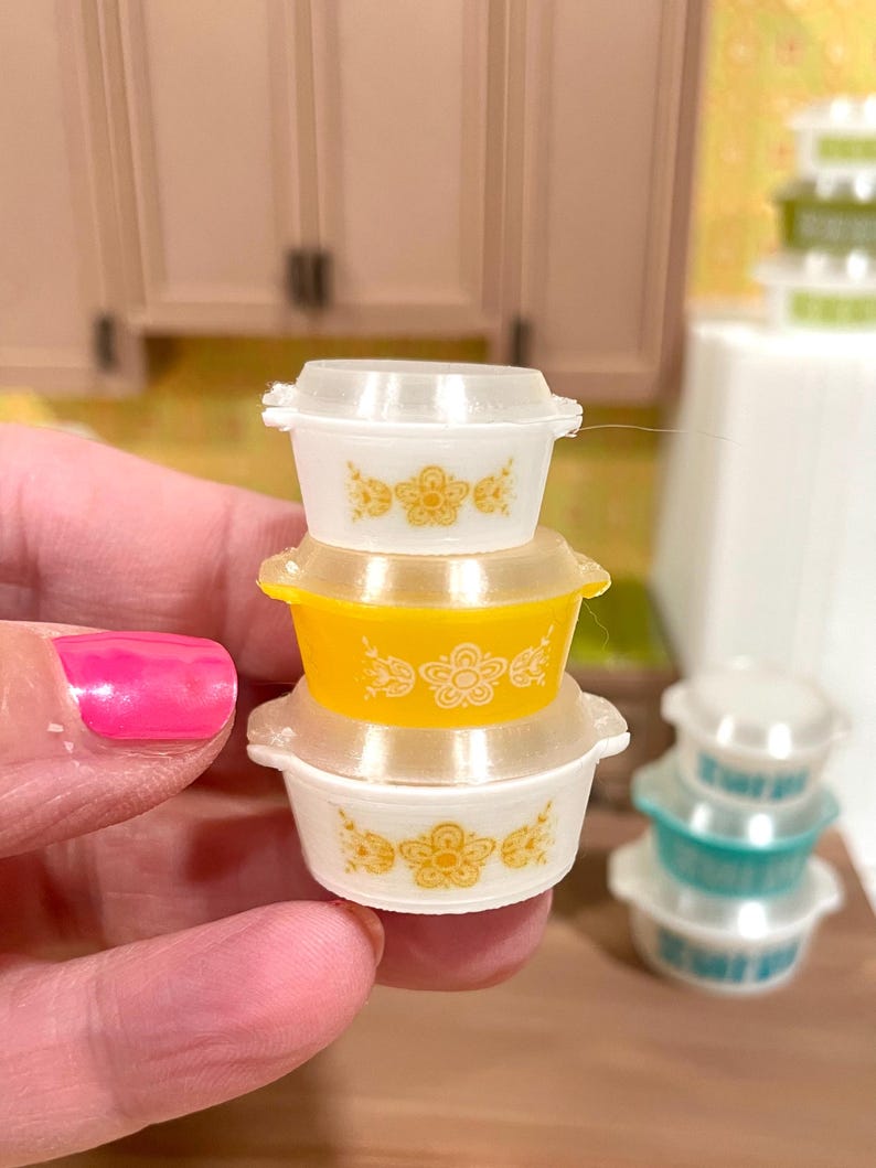 May include: A stack of miniature Pyrex-style bowls. The stack includes a yellow bowl, two white bowls, and clear lids. Each bowl features a floral design in yellow. Other bowls are visible in the background.