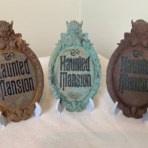 May include: Three Haunted Mansion signs with a gargoyle on top. The signs are made of a dark brown material and have a distressed look. The text on the signs reads "The Haunted Mansion".
