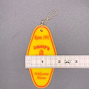 May include: Yellow and red plastic key chain shaped like a hotel room key with the text "Room 1991" and "Disney's" with a small image of Cinderella's castle. The key chain also has the text "Welcome Home".