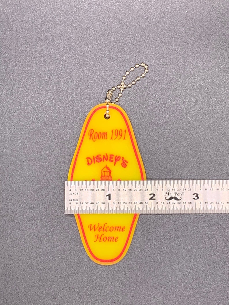 May include: Yellow and red plastic key chain shaped like a hotel room key with the text "Room 1991" and "Disney's" with a small image of Cinderella's castle. The key chain also has the text "Welcome Home".