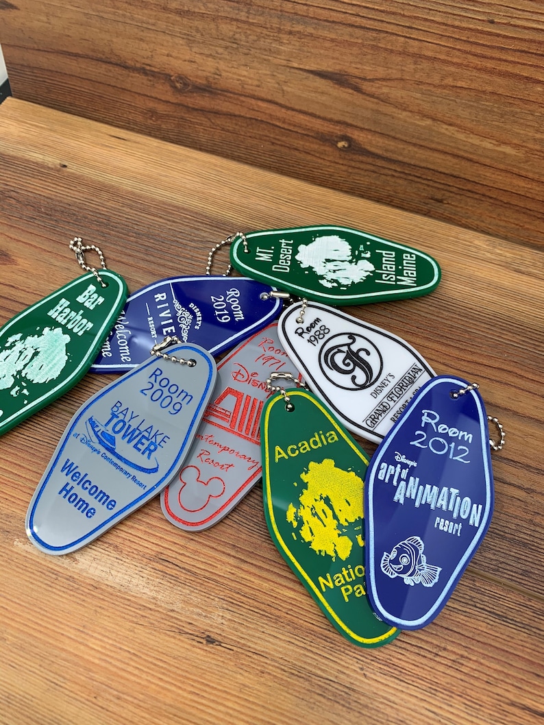 May include: A collection of colorful keychains shaped like hotel room keys. Each keychain features a different Disney resort, including Bay Lake Tower, the Contemporary Resort, Disney's Grand Floridian Resort & Spa, and Disney's Art of Animation Resort. The keychains are made of metal and have a shiny finish.