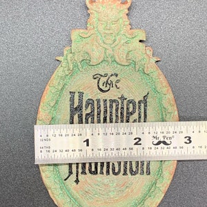 May include: A green and copper-colored metal sign with a gargoyle at the top and the words "The Haunted Mansion" in black lettering. The sign is approximately 7 inches in diameter.