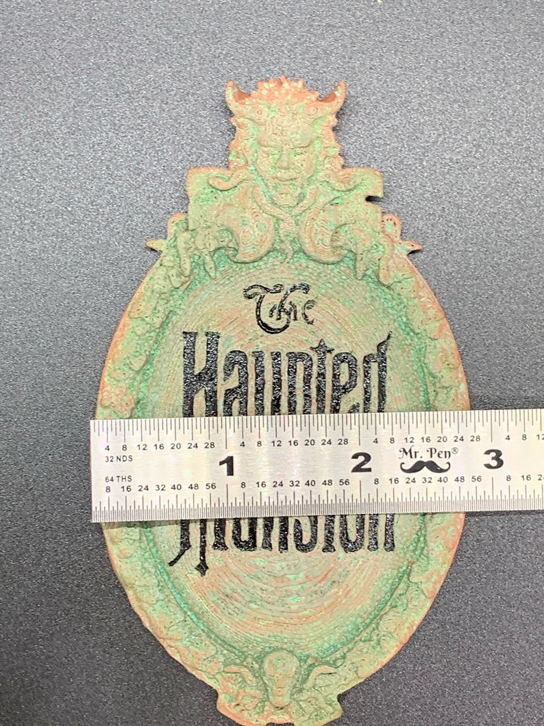 May include: A green and copper-colored metal sign with a gargoyle at the top and the words "The Haunted Mansion" in black lettering. The sign is approximately 7 inches in diameter.
