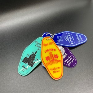 May include: A collection of five vintage Disney hotel keychains. The keychains are shaped like hotel room keys and feature different Disney resorts, including Cinderella's Castle, Old Key West, and the Riviera. The keychains are made of plastic and have a metallic finish. The keychains are attached to a silver chain.