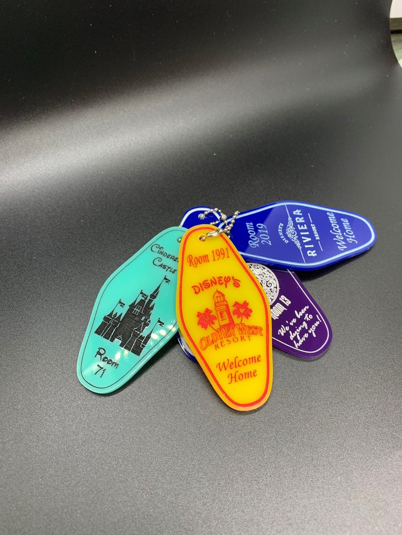 May include: A collection of five vintage Disney hotel keychains. The keychains are shaped like hotel room keys and feature different Disney resorts, including Cinderella's Castle, Old Key West, and the Riviera. The keychains are made of plastic and have a metallic finish. The keychains are attached to a silver chain.