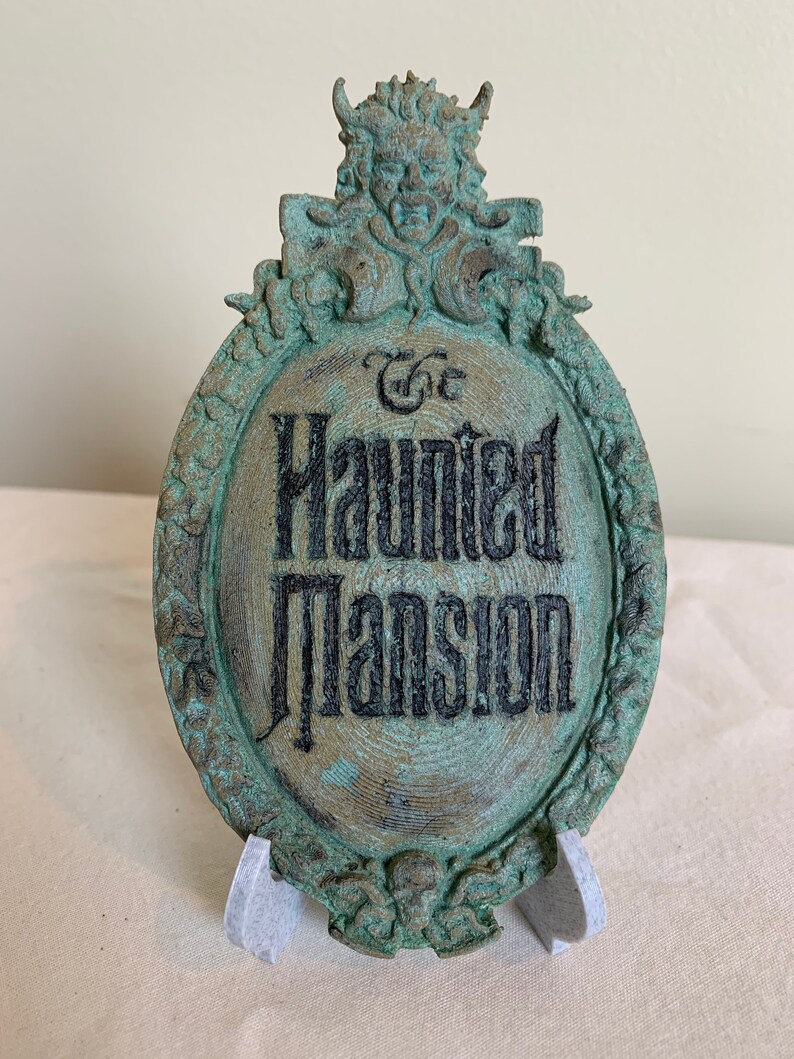 May include: A green metal sign with a skull and floral design. The sign reads "The Haunted Mansion".