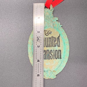 May include: A green and gold-colored metal ornament with the text "The Haunted Mansion" in black lettering. The ornament has a red ribbon attached to the top.