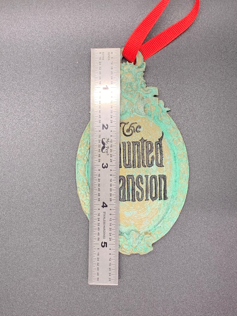 May include: A green and gold-colored metal ornament with the text "The Haunted Mansion" in black lettering. The ornament has a red ribbon attached to the top.