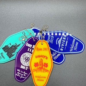 May include: A collection of five colorful motel keychains. The keychains are shaped like a traditional motel key with rounded edges. The keychains are purple, blue, yellow, and turquoise. The keychains have text on them, including "Room 1991", "Disney's", "Old Key West Resort", "Welcome Home", and "Gracey Manor B&B".