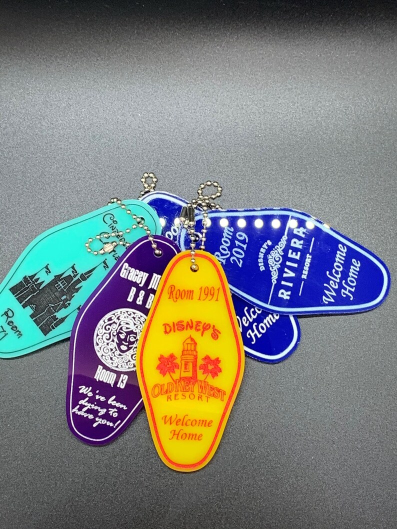 May include: A collection of five colorful motel keychains. The keychains are shaped like a traditional motel key with rounded edges. The keychains are purple, blue, yellow, and turquoise. The keychains have text on them, including "Room 1991", "Disney's", "Old Key West Resort", "Welcome Home", and "Gracey Manor B&B".