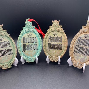May include: Four green and brown metal ornaments with a distressed finish. Each ornament features a sculpted image of a ghostly figure and the text "The Haunted Mansion".