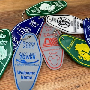 May include: A collection of colorful Disney resort hotel keychains. The keychains are shaped like hotel room keys and feature the names of the resorts, including Bay Lake Tower, Contemporary Resort, Grand Floridian Resort & Spa, and Acadia. The keychains are made of plastic and have a silver metal ring.