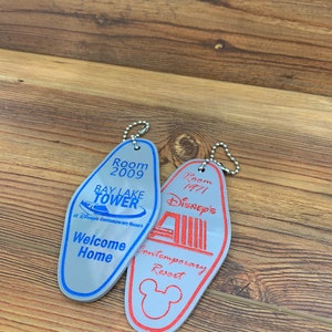 May include: Two silver keychains shaped like hotel room keys. The keychain on the left is blue and says "Room 2009 Bay Lake Tower at Disney's Contemporary Resort" and "Welcome Home". The keychain on the right is red and says "Room 1971 Disney's Contemporary Resort".