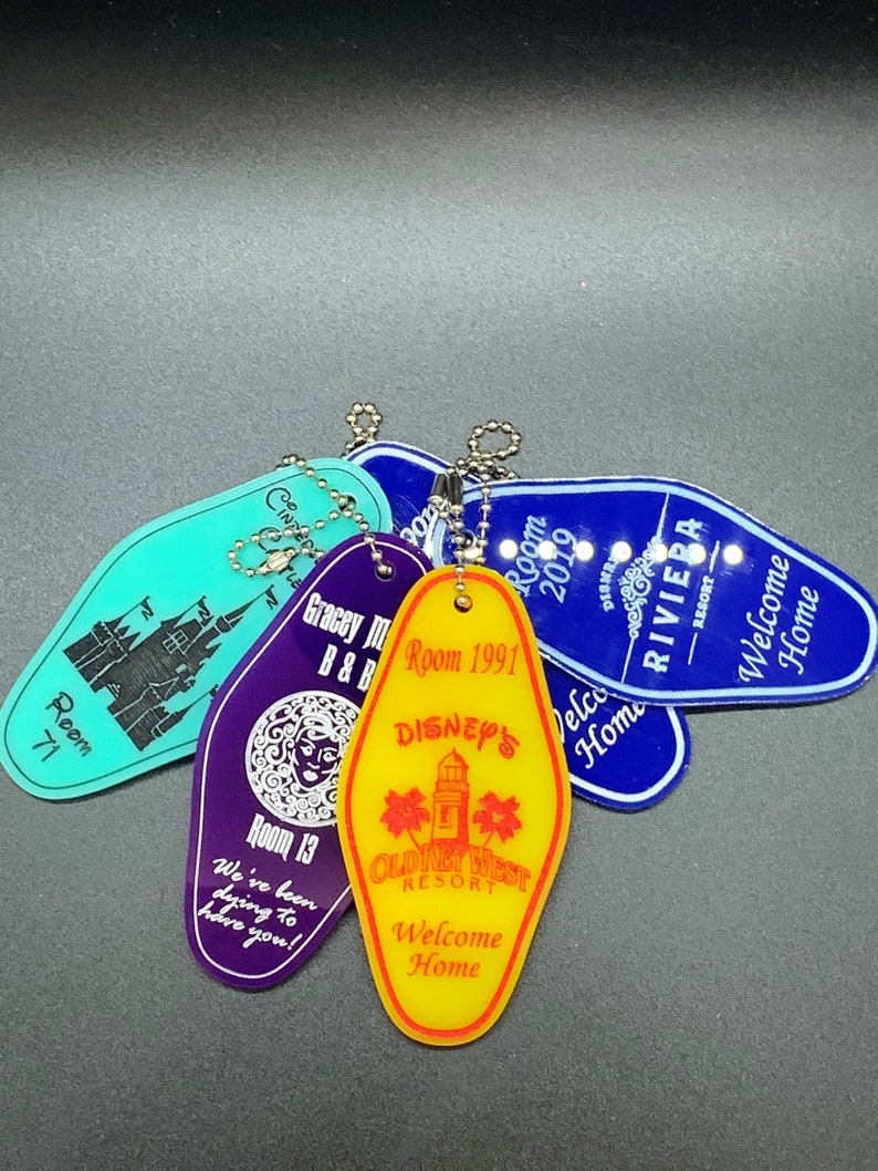 May include: A collection of five colorful keychains shaped like motel room keys. Each keychain features a different Disney-themed design, including a castle, a mermaid, a lighthouse, and a western town. The keychains are made of plastic and have a silver metal ring.
