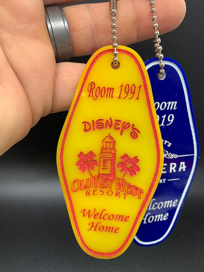 May include: Yellow and blue keychains with a red inscription. The yellow keychain has a lighthouse, palm trees, and the text "Disney's Old Key West Resort Welcome Home". The blue keychain has the text "Room 19".