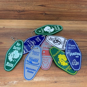 May include: A collection of colorful keychains with various designs, including a green keychain with the text "Bar Harbor Maine", a blue keychain with the text "Room 2012 Disney's Art of Animation Resort", and a green keychain with the text "Acadia National Park".