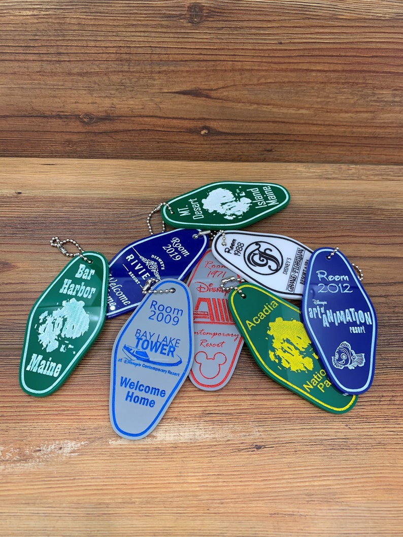 May include: A collection of colorful keychains with various designs, including a green keychain with the text "Bar Harbor Maine", a blue keychain with the text "Room 2012 Disney's Art of Animation Resort", and a green keychain with the text "Acadia National Park".
