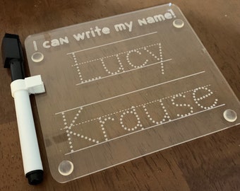 May include: A clear plastic dry-erase board with the text "I can write my name!" at the top. The board has two lines with dotted letters for the names "Lucy Krause". A white dry-erase marker is next to the board.