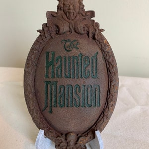 May include: A brown, weathered, oval-shaped sign with a green inscription that reads "The Haunted Mansion". The sign is topped with a sculpted gargoyle with horns and a menacing expression.