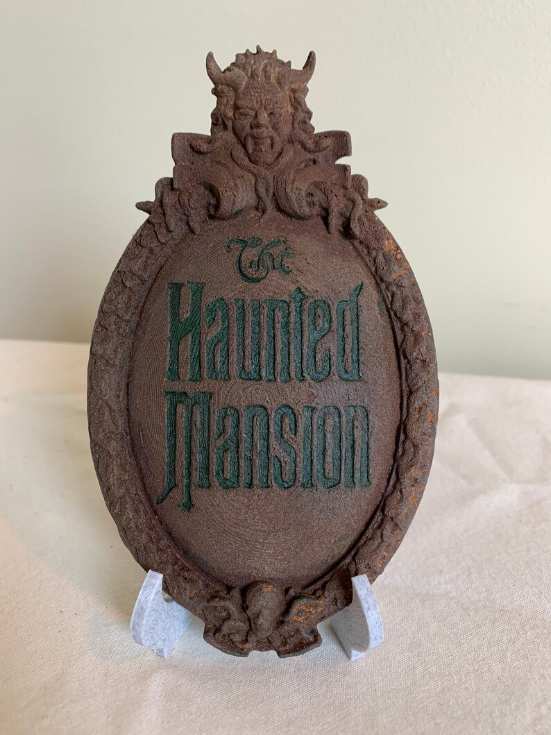 May include: A brown, weathered, oval-shaped sign with a green inscription that reads "The Haunted Mansion". The sign is topped with a sculpted gargoyle with horns and a menacing expression.