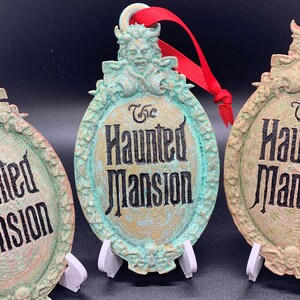 May include: Three green and gold Haunted Mansion Christmas ornaments with a red ribbon. The ornaments feature a vintage-style design with the words "The Haunted Mansion" in black lettering.