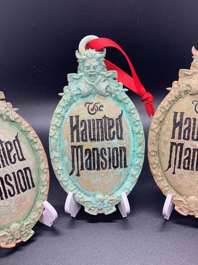 May include: Three green and gold Haunted Mansion Christmas ornaments with a red ribbon. The ornaments feature a vintage-style design with the words "The Haunted Mansion" in black lettering.