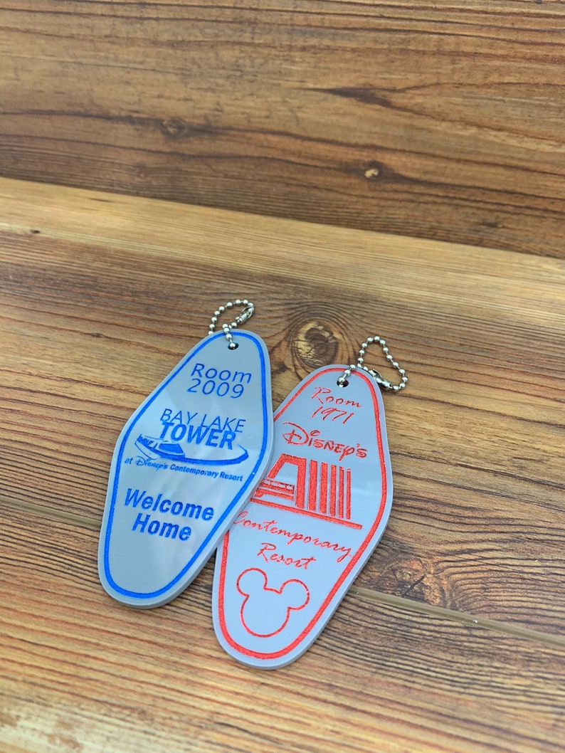 May include: Two silver keychains shaped like hotel room keys. The keychain on the left is blue and says "Room 2009 Bay Lake Tower at Disney's Contemporary Resort" and "Welcome Home". The keychain on the right is red and says "Room 1971 Disney's Contemporary Resort".