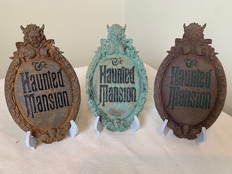 May include: Three Haunted Mansion signs with a gargoyle on top. The signs are made of a dark brown material and have a distressed look. The text on the signs reads "The Haunted Mansion".