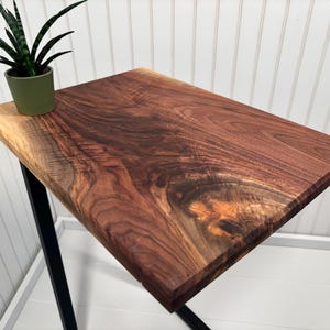 May include: A rectangular side table featuring a natural live edge walnut wood top with visible dark brown grain patterns and a knot, supported by a black metal frame. A small green potted plant sits on the corner of the wooden surface.