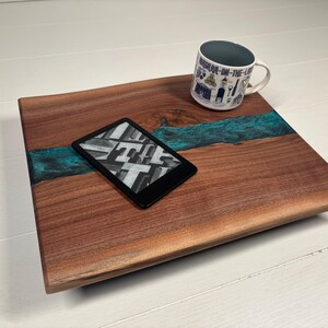 May include: A rectangular wooden board with a natural grain and a central turquoise resin inlay. A black e-reader displaying a grid of letters rests on the surface, accompanied by a white ceramic mug featuring blue illustrations and the text Niagara-on-the-Lake.